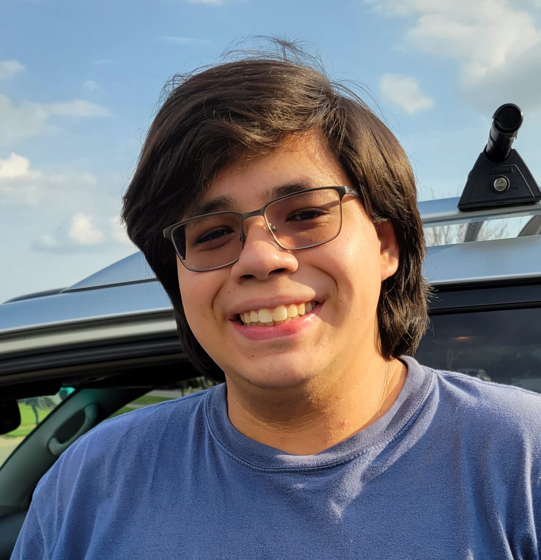 Photo of a young Indigenous man in a blue t-shirt and glasses with a side part in short hair, smiling for the camera