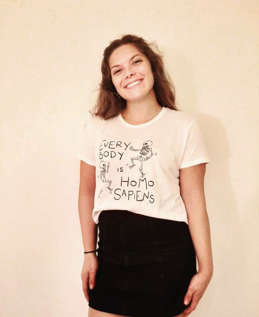 Image of a white woman with shoulder length brown hair and a nose ring, wearing a white t-shirt with cartoon skeletons that says "every body is Homo sapiens" and a short black skirt