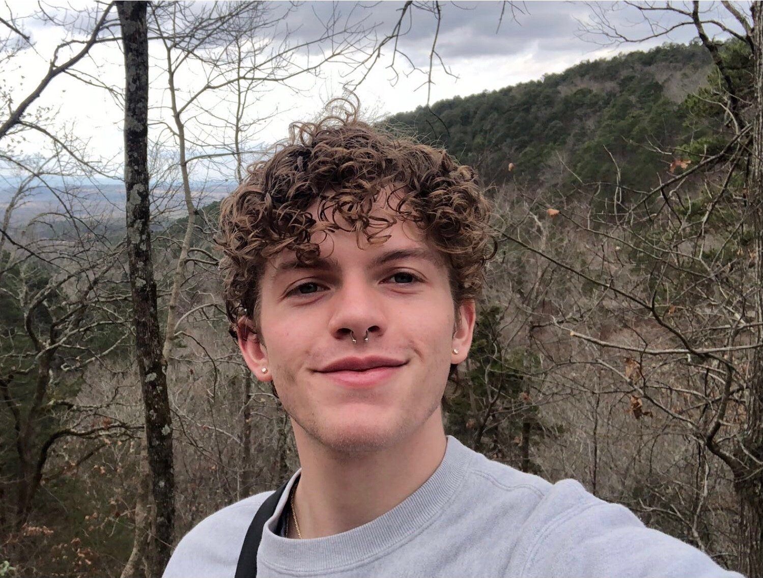 selfie of a young white man with short curly brown hair falling over his forehead, with a septum piercing and studs in each earlobe, wearing a gray tshirt in a wooded area