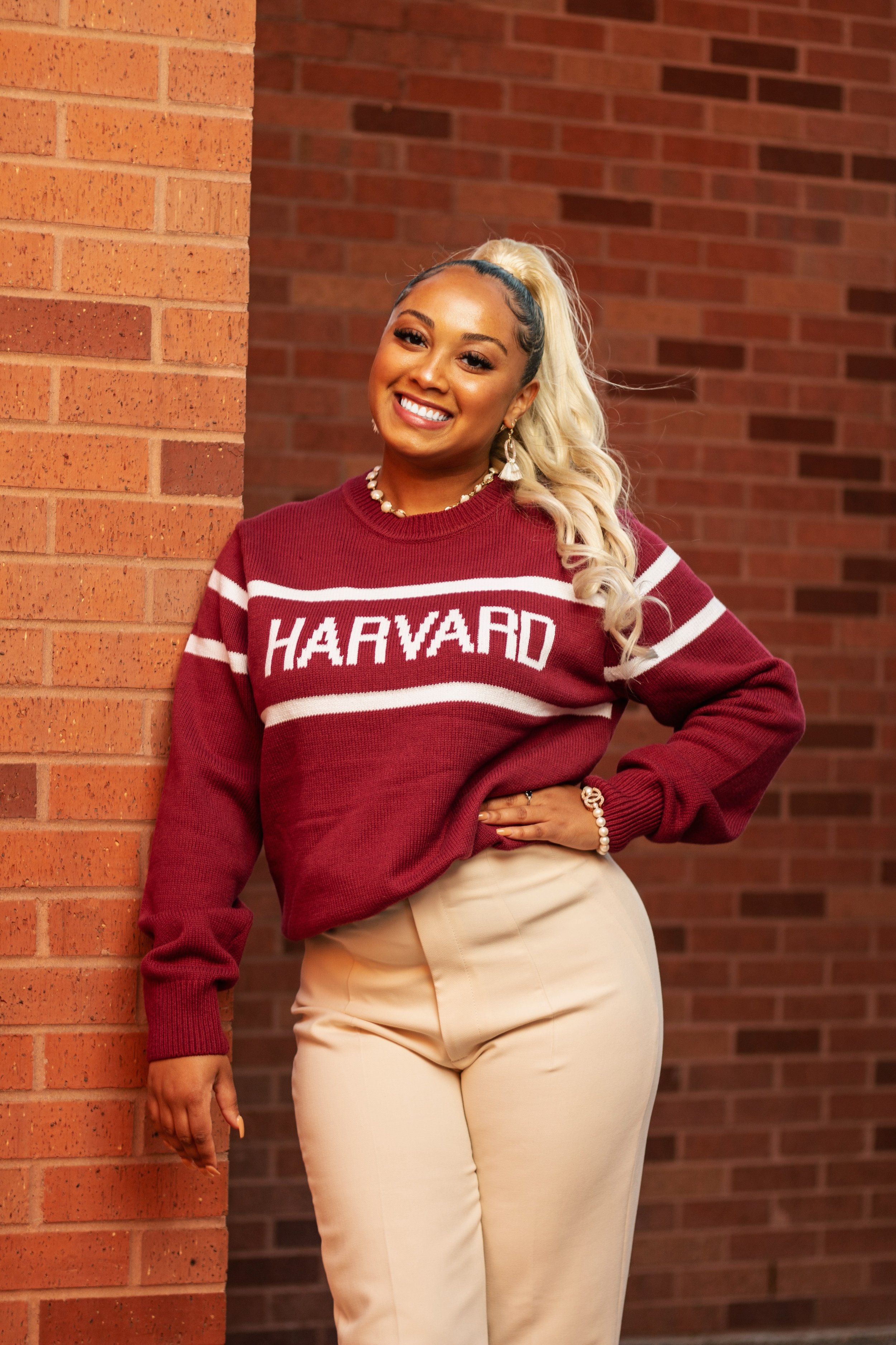 Photo of a Black woman smiling and leaning against a brick wall, smiling at the camera with her hair in a blond ponytail, wearing tan pants and a knit sweater that says Harvard